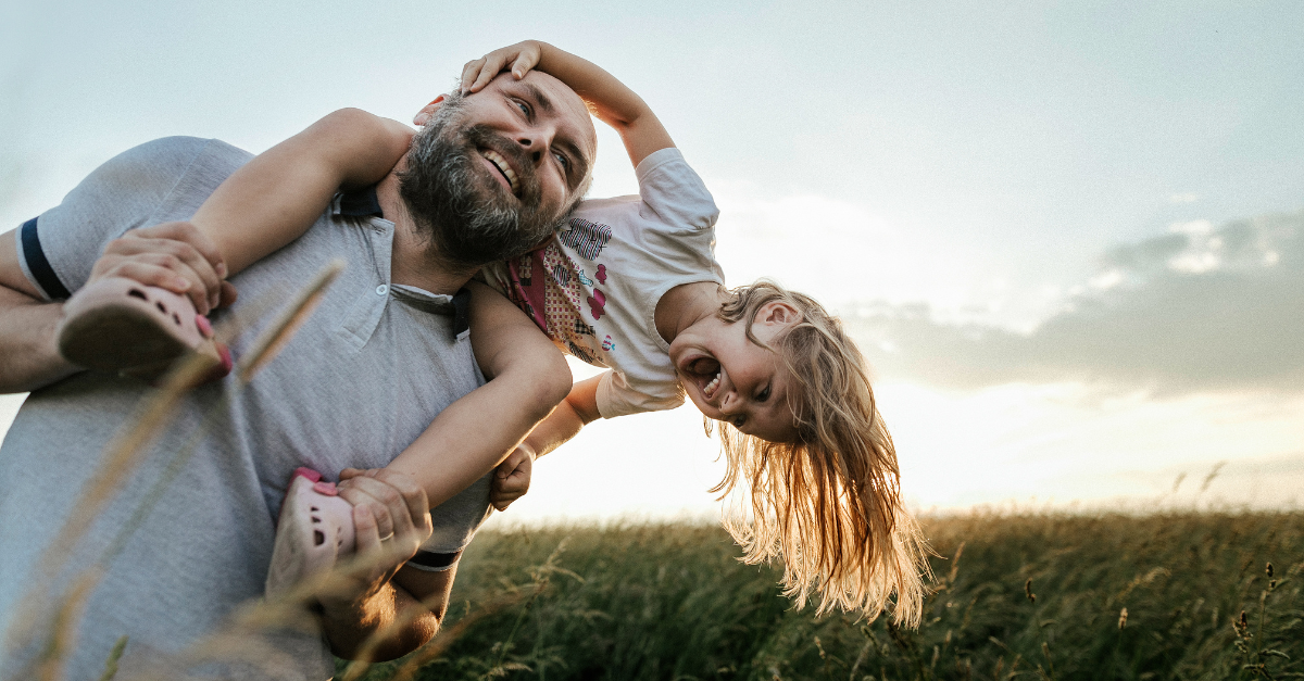 A child rides on an adult's shoulders as they walk through a green field. Both are laughing. 